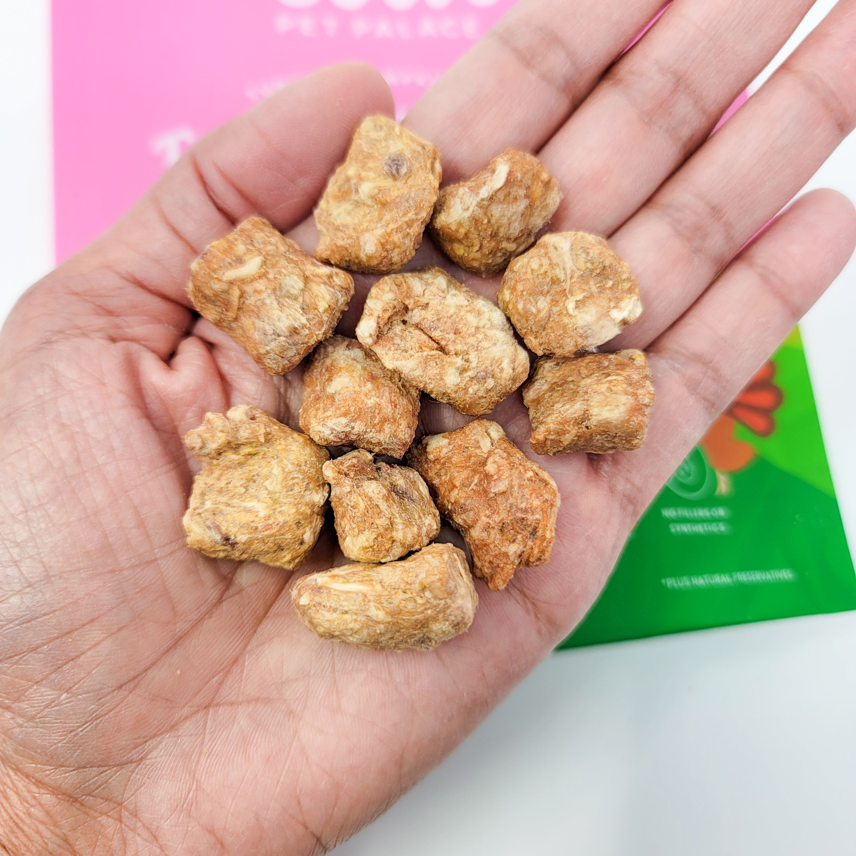 Hand holding a small pile of brown pet treats with a pink and green box in the background.